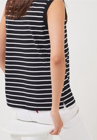 Zwart-wit gestreepte mouwloze top van zacht materiaal, met een ronde onderkant en zijsplitten, voorzien van een klein rood logo label.