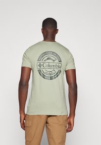 Lys grønn bomull-T-skjorte med et sirkulært grafisk design på ryggen, som har Columbia-logoen, fjell og skogmønstre.