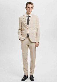 Beige kostym i slät tyg med en figursydd design. Har en tvåknappparad stängning, notch-revers och smala byxor. Kombineras med ett svart slips.