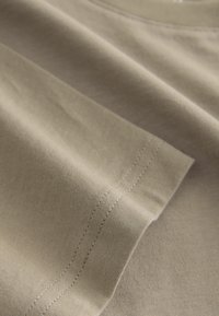 Beige színű pamut póló kerek nyakkivágással, amely lágy textúrával és jól látható varrási részletekkel rendelkezik az ujjújaknál és az alján.