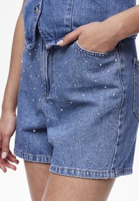 Close-up van een persoon die blauwe denim shorts met zilveren studs draagt, met één hand in de zak, gecombineerd met een bijpassende blauwe denim top.