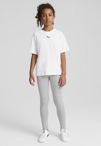 Fehér, rövid ujjú póló fekete logóval, szürke, testhezálló leggings-szel és fehér sportcipővel, semleges háttér előtt.