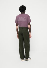 Camiseta gráfica color lila con el texto "OBEY STUDIOS", combinada con pantalones verdes oliva. Los pantalones cuentan con un bolsillo trasero y son ligeramente holgados.