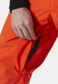 Heldere oranje waterdichte broek heeft een zijzak met rits en zwarte gaasvoering. De stof is glad met subtiele stikdetails.