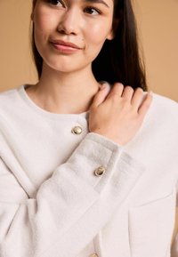 Weiße Wolljacke mit runder Ausschnitt, geknöpften Manschetten und frontalen Taschen mit goldfarbenen Beschlägen. Weicher, strukturierter Stoff.