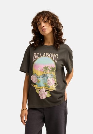 Kobieta z kręconymi włosami, ubrana w ciemny t-shirt Billabong z tropikalnym zachodem słońca, palmami, różowymi kwiatami i tekstem "Dreams of Paradise".