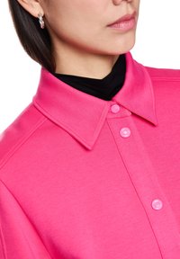 Rosa Hemd mit Kragen aus weichem Stoff, mit Knopfverschluss und glatter Textur, über einem schwarzen Kleidungsstück getragen.