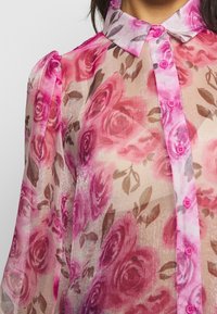 Gennemsigtig bluse med pink rosemønster, knappet fortil og en blød krave. Har pink knapper og let puffede ærmer.
