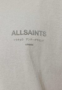 Gaiši pelēks T-kreklis ar "ALLSAINTS" logotipu, papildus teksts japāņu valodā un vārds "LONDON" zem tā, izdrukāts mīkstākā pelēkā krāsā uz auduma.