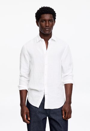 Un homme aux cheveux courts et bouclés portant une chemise blanche à manches longues boutonnée et un pantalon sombre, debout devant un fond blanc uni.