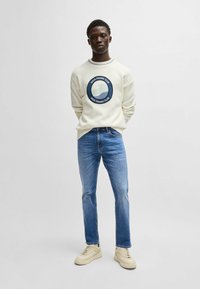 Suéter beige con un logo azul circular, combinado con jeans rectos de un azul claro y zapatillas beige. Diseño simple y casual.