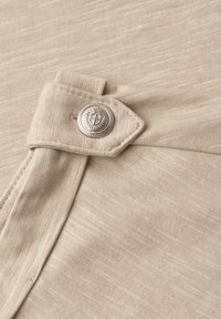 Beige fabric with a stitched flap fastened by a round silver button embossed with a crest and laurel wreath design.