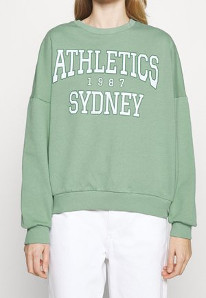 Lichtgroene trui met witte tekst "ATHLETICS 1987 SYDNEY", gedragen door een persoon met een witte broek.