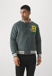 Giacca bomber in corduroy verde scuro, con logo ricamato "B" giallo e polsini e orlo a coste bianchi. Chiusura frontale con bottoni.