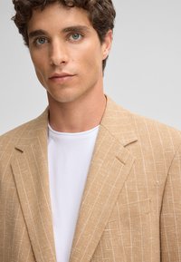Beiger Blazer aus strukturiertem Stoff, mit dezentem weißen Streifenmuster, Reverskragen und einer einzelnen Brusttasche. Darunter ein weißes T-Shirt mit Rundhalsausschnitt.