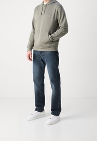 Sudadera con capucha en tejido verde apagado con bolsillo tipo canguro y parche con logo, combinada con vaqueros azul oscuro y zapatillas blancas.