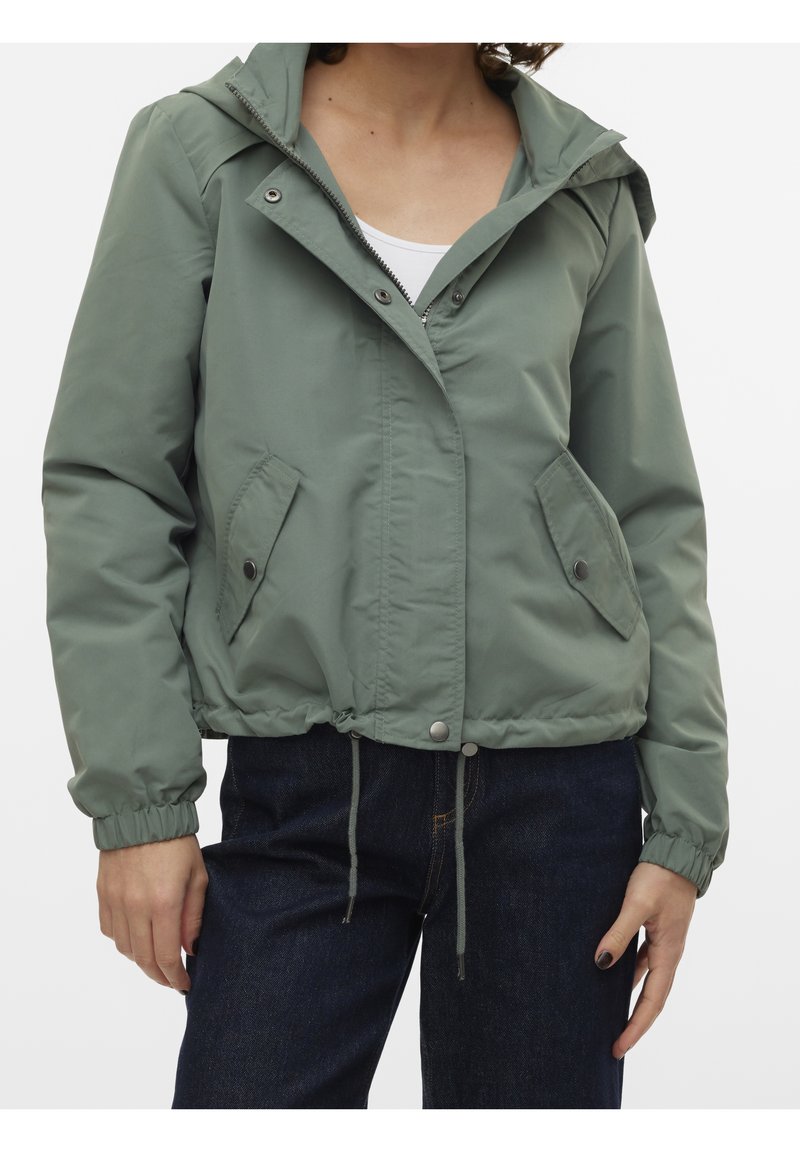 Personne portant une veste coupe-vent verte à capuche avec poches pressionnées et poignets élastiques sur un jean foncé et un haut blanc.