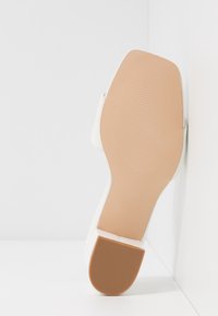 Semelle d'une chaussure à plat blanche avec un dessous texturé beige et un talon en caoutchouc, positionnée verticalement contre un coin blanc.