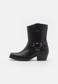 Botas de tobillo de cuero negro con punta redonda, tacón apilado y una correa ancha con herrajes plateados y un detalle circular.