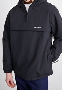 Hombre que lleva una chaqueta con capucha de Carhartt negra, con cremallera frontal y gran bolsillo en el pecho, combinada con jeans azul oscuro.
