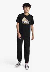 Μαύρο μπλουζάκι με γραφικό απεικόνισης ενός καφέ sneaker Converse, συνδυασμένο με μαύρη φόρμα και μαύρα και λευκά αθλητικά παπούτσια. Απλός σχεδιασμός.