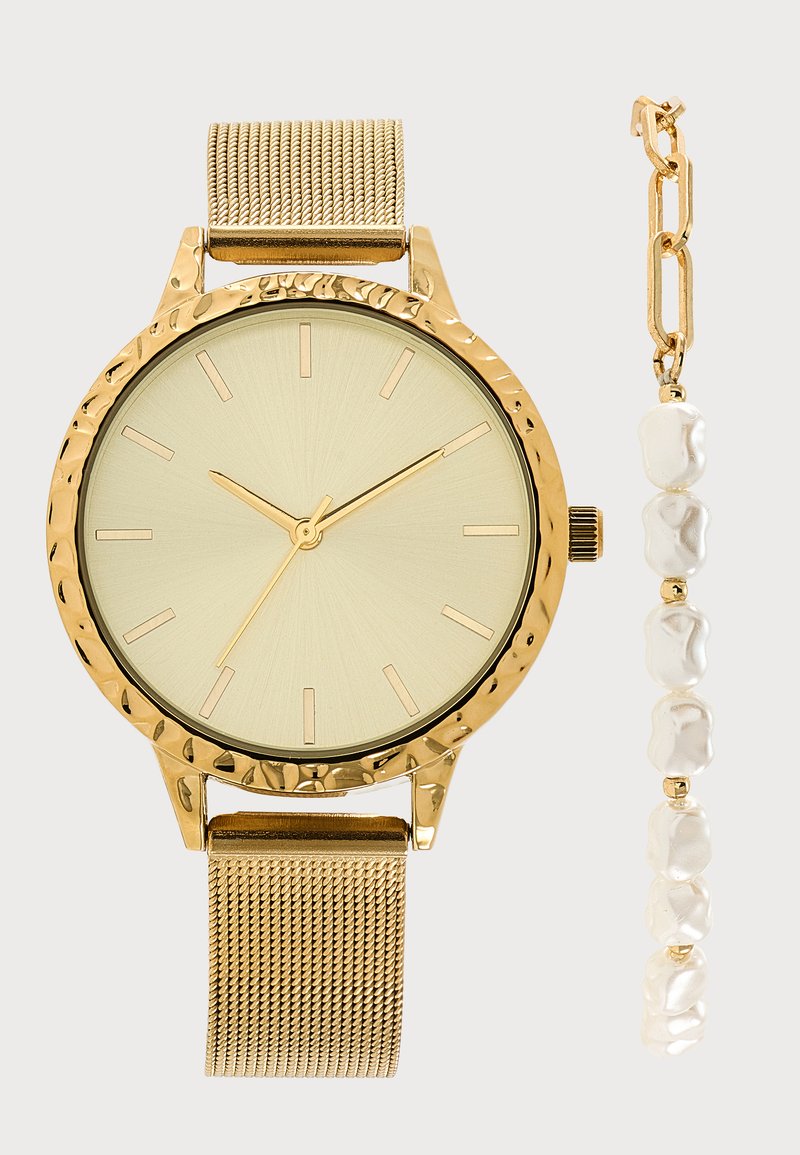 Χρυσό ρολόι χειρός με υφή στην στεφάνη και πλεγμένο λουράκι, τοποθετημένο δίπλα σε χρυσό βραχιόλι αλυσίδα με ακανόνιστα λευκά χάντρες.