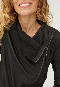 Leende person med lockigt blont hår som bär en svart jacka med en asymmetrisk krage och en stor diagonal dragkedja.