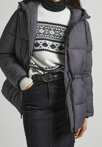 Šedá prošívaná bunda s kapucí, vybavená zipem a elastickými manžetami, vrstvená přes vzorovaný šedý svetr a černou sukni, stažená černým páskem.