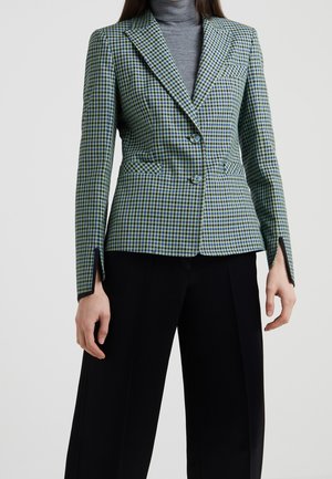 Frau trägt einen grün-blau karierten Blazer über einem grauen Rollkragenpullover mit schwarzen weit geschnittenen Hosen und steht vor einem schlichten Hintergrund.