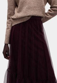 Osoba w beżowym dzianinowym swetrze wpuszczonym w ciemnofioletową spódnicę z tiulu, z jedną ręką opartą na biodrze, a drugą opuszczoną wzdłuż ciała.