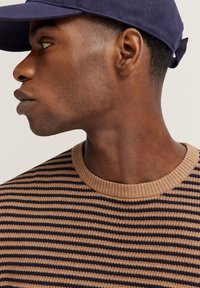 Gestreifter Strickpullover in Braun und Marineblau mit einem gerippten Rundhalsausschnitt. Dunkelblaues BaseballCap teilweise sichtbar.