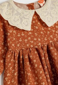 Ruskea kukkakuosiset mekko, jossa on teksturoitu kangas, vaaleanvärinen brodeerattu kaulus ja kootut hameen yksityiskohdat.