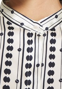 Chemise en tissu blanc avec des rayures noires verticales et des motifs circulaires. Comprend un col montant et une patte de boutonnage. Texture lisse.
