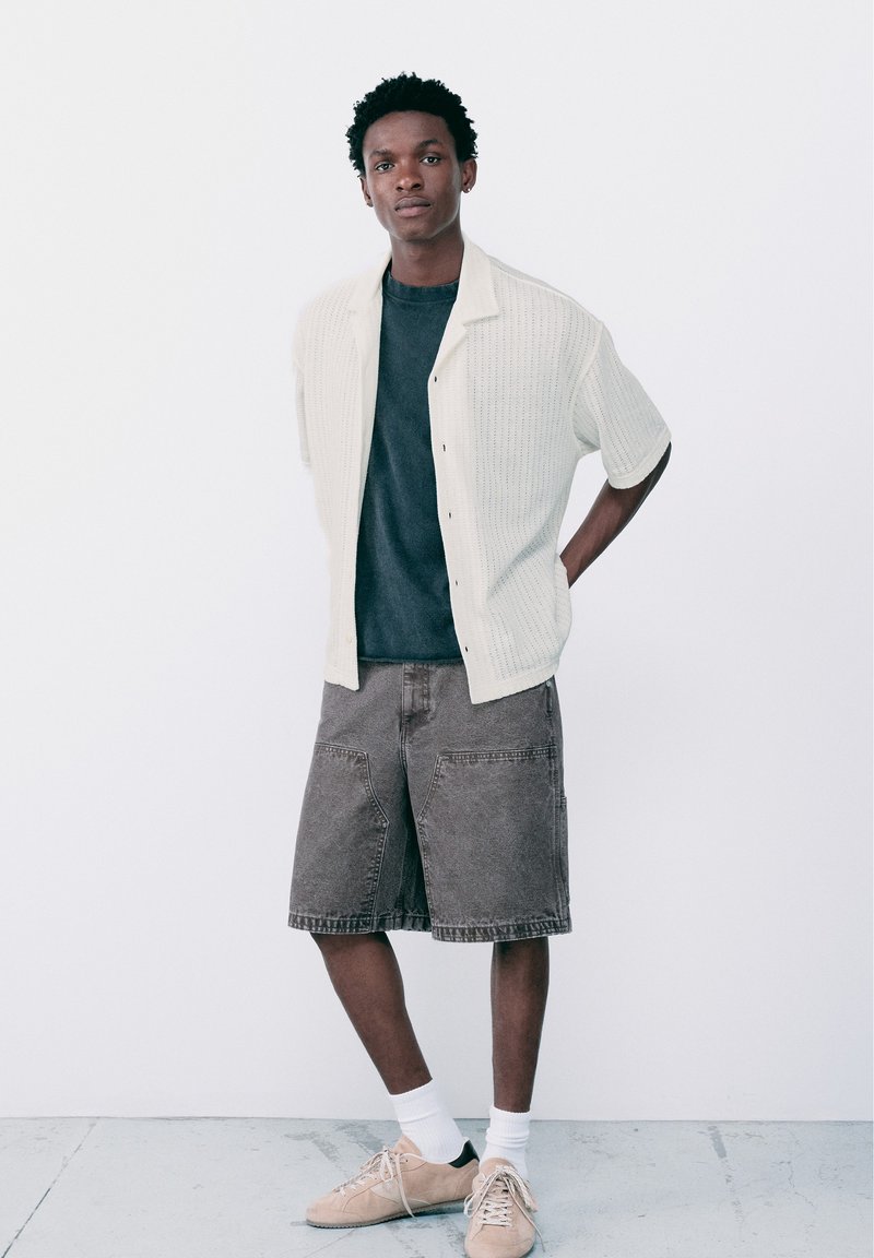 Un jeune homme se tient contre un mur blanc, portant une chemise blanche à manches courtes en maille ouverte, un t-shirt foncé, un short gris au niveau des genoux, des chaussettes blanches et des baskets beiges.