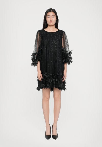 Crna haljina s cvjetnim mrežastim slojem, napučenim rukavima i volanima na rubu. Uparena s crnim cipelama s oštrim vrhovima.