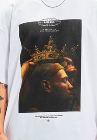 Hvid bomulds T-shirt med et stort, tekstureret grafisk motiv af en kronet figur med gyldne accenter, samt et tekstcitat nedenfor.
