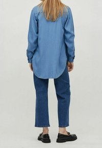 Chemise en denim bleu clair à manches longues et ourlet arrondi, associée à un jean bleu foncé. Portée avec des chaussures plates noires. Vue de dos.