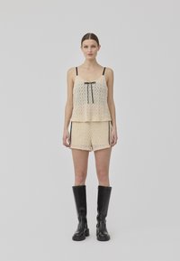 Spitzen-Camisole mit schwarzen Trägern, die eine Schleife zeigt. Kombiniert mit passenden Shorts, beide in einem hellen Beige mit einem strukturierten Muster. Schwarze knielange Stiefel.