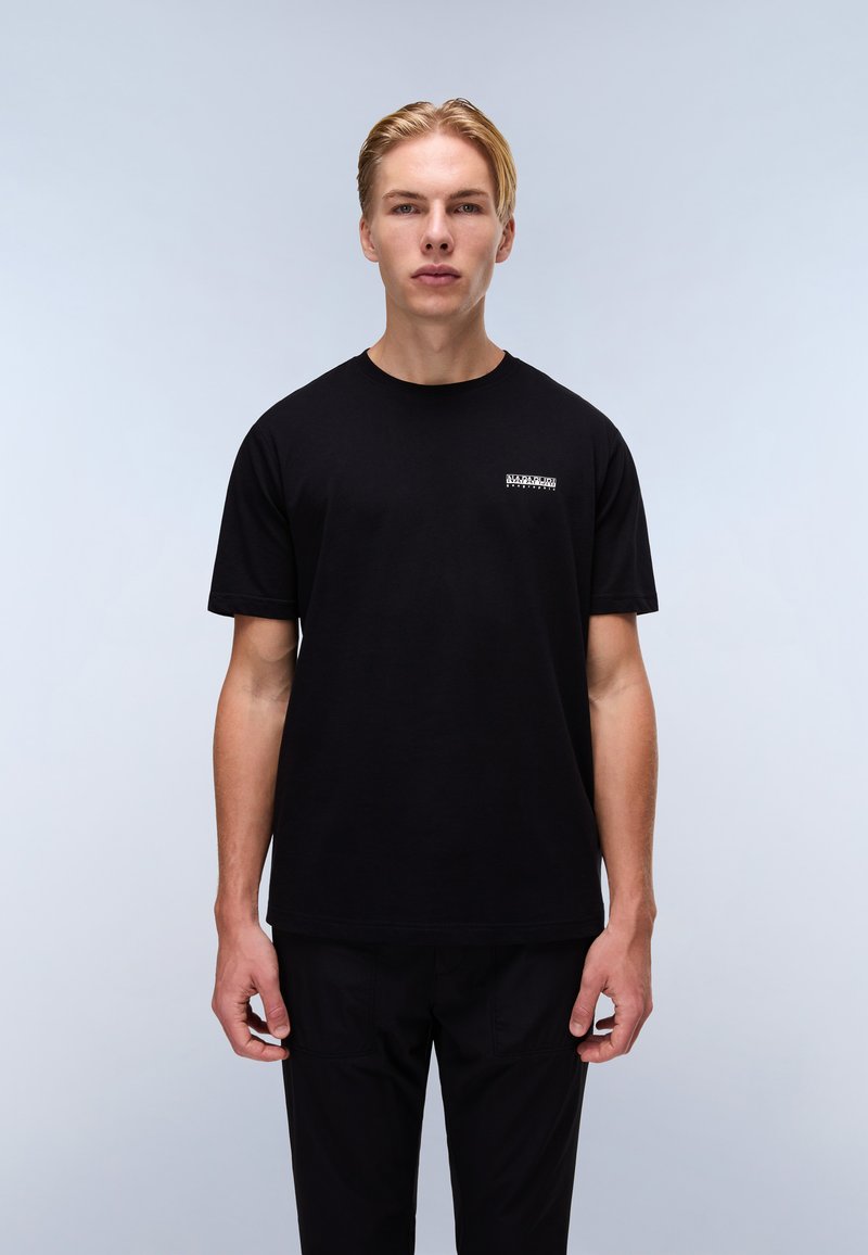 T-shirt nera in cotone con maniche corte, scollo rotondo e una piccola grafica sulla parte sinistra del petto. Modello che indossa pantaloni neri su uno sfondo chiaro.