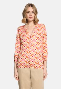 Femme aux cheveux ondulés mi-longs portant un chemisier à manches longues avec un motif géométrique multicolore et un pantalon beige, sur fond blanc.