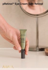 Crème pour les yeux pRetinol™ d'AHAVA dans un tube vert, avec un bouchon noir à vis. Placée sur une surface claire, tenue par une main. Miroir circulaire en arrière-plan.