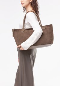 Borsa tote in suede marrone con due manici, caratterizzata da un'esterno morbido e dettagli hardware sottili. Tenuta da una persona che indossa un maglione bianco.