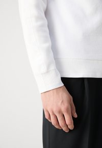 Højre hånd afslappet ved siden, iført en hvid langærmet trøje med teksturerede ærmer og sorte bukser mod en lys baggrund.