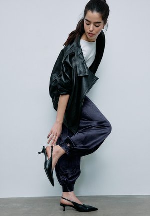 Donna con giacca di pelle nera, top bianco e pantaloni jogger in satin scuro che sistema una scarpa nera con tacco basso a cinturino sul pavimento grigio e sfondo a parete semplice.