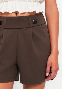 Shorts taillés marron avec deux plis avant et deux boutons noirs à la taille. Texture lisse; l'ourlet arrive à mi-cuisse.