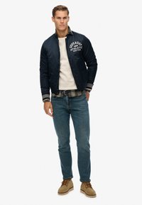 Chubasquero navy con un logo blanco, suéter crema, camisa a cuadros, vaqueros azules y botas beige, con puños a rayas en contraste.