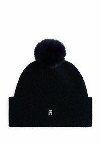 Marineblauwe wollen beanie met een grote bonten pompon bovenop en een omgeslagen manchet met een gouden logo-accent. De textuur is gebreid en dicht.