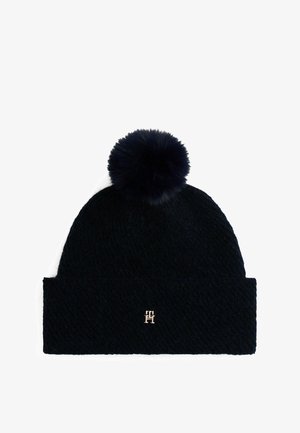 Gorro de lana navy con un gran pompón de piel en la parte superior y un dobladillo que presenta un acento de logo dorado. La textura es de punto y densa.