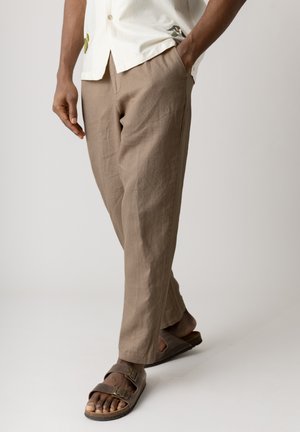 Mand iført beige bukser med lige ben, hvid kortærmet skjorte og brune sandaler med to remme, stående med den ene hånd i lommen.