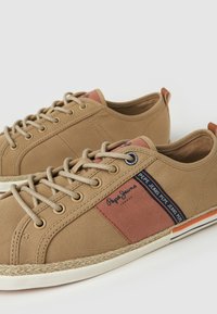 Zapatillas de lona beige con acentos marrones, cordones redondos, suela a rayas y parche de marca. Presentan una superficie texturizada y un panel de color contrastante.